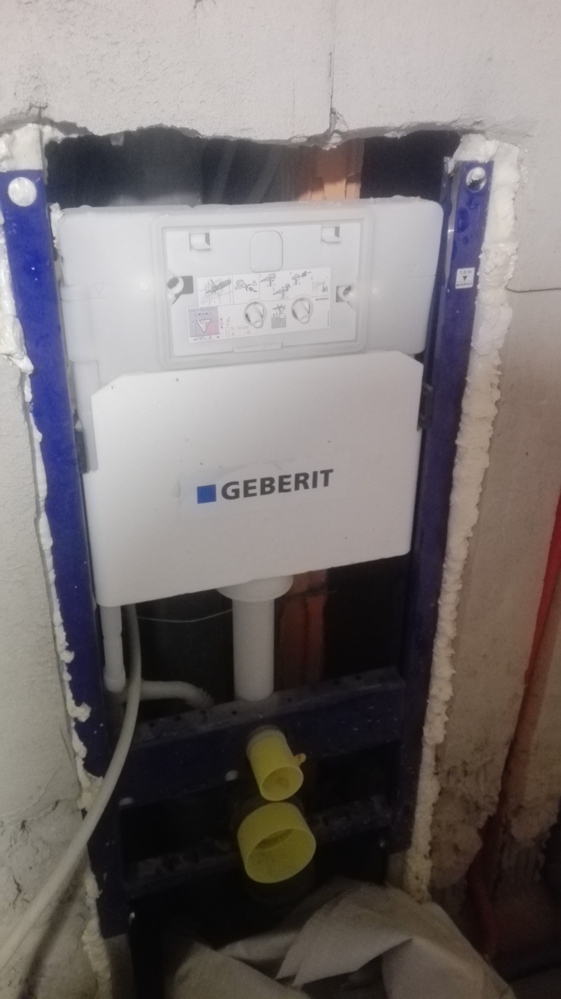 Stelaż podtynkowy Geberit zamontowany w ścianie, widoczne przyłącza. Prace instalacyjne w łazience w Poznaniu, przed zabudową. Niebieska rama i biały zbiornik.