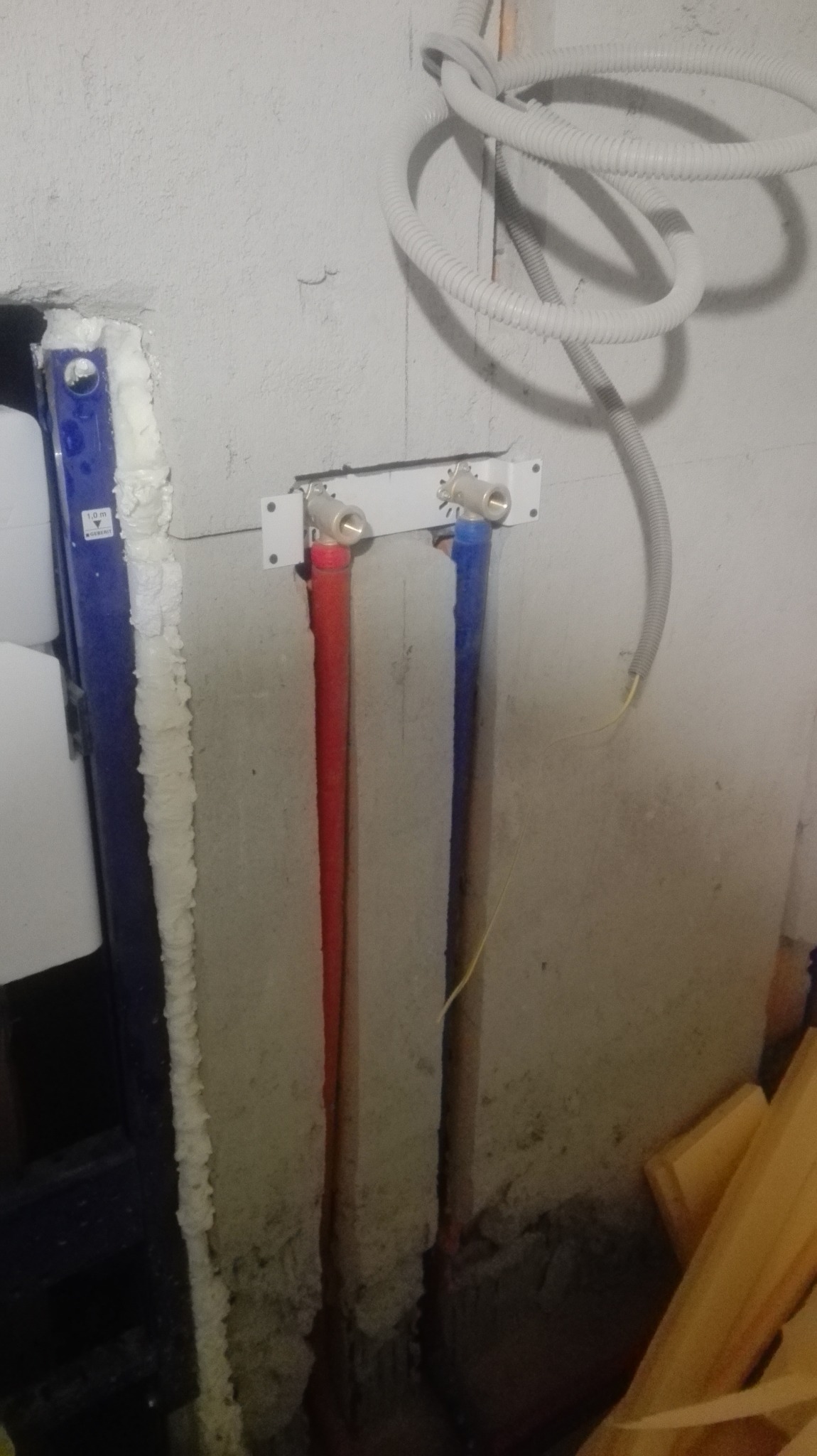 Instalacja wodna w łazience: czerwona i niebieska rura doprowadzona do zaworów, widoczne przewody elektryczne i stelaż podtynkowy. Surowy stan, przygotowanie do wykończenia.