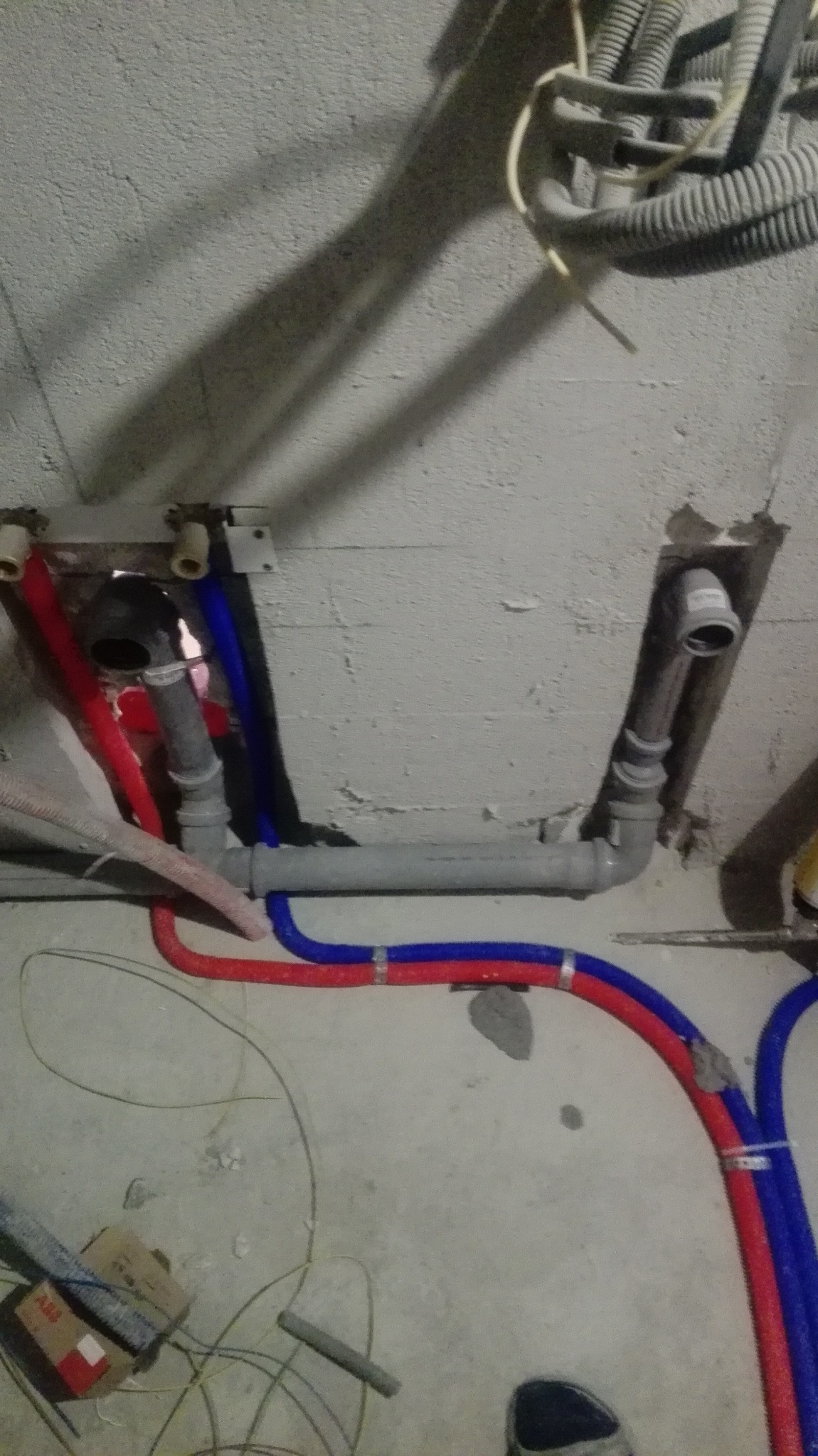 Instalacja wodno-kanalizacyjna w trakcie montażu: rury czerwone i niebieskie, szare rury kanalizacyjne, widoczne mocowania i fragment surowego sufitu.