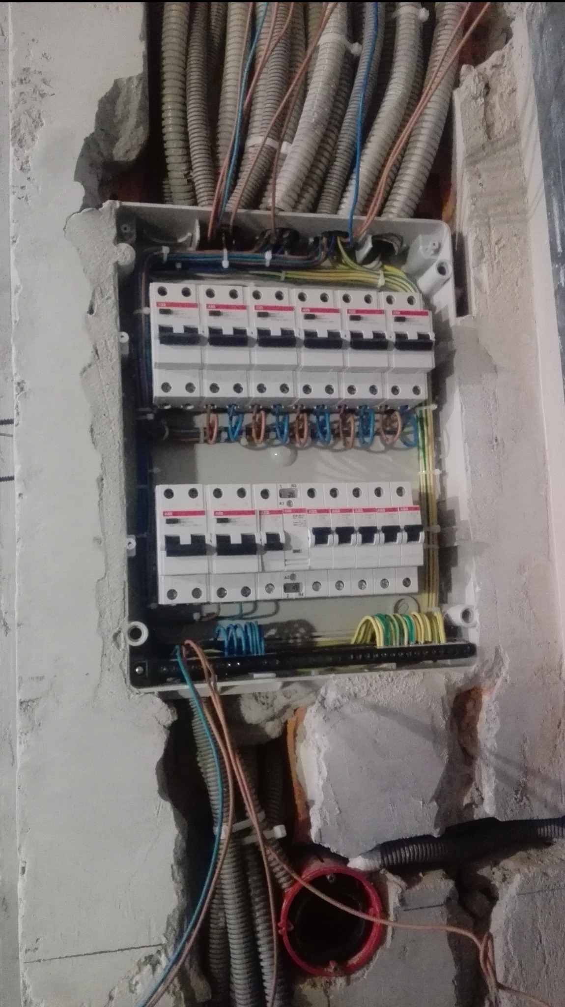 Rozdzielnica elektryczna z przewodami w ścianie, widoczne zabezpieczenia i kable w peszlach. Instalacja w trakcie prac, surowy stan ściany.