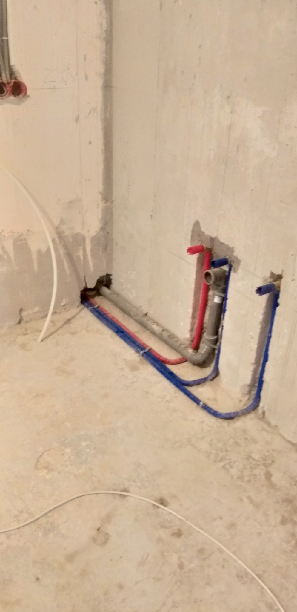 Surowy kąt łazienki z instalacją wodną: rury miedziane i plastikowe w kolorach czerwonym i niebieskim, wystające ze ściany. Widoczne ślady po tynku.