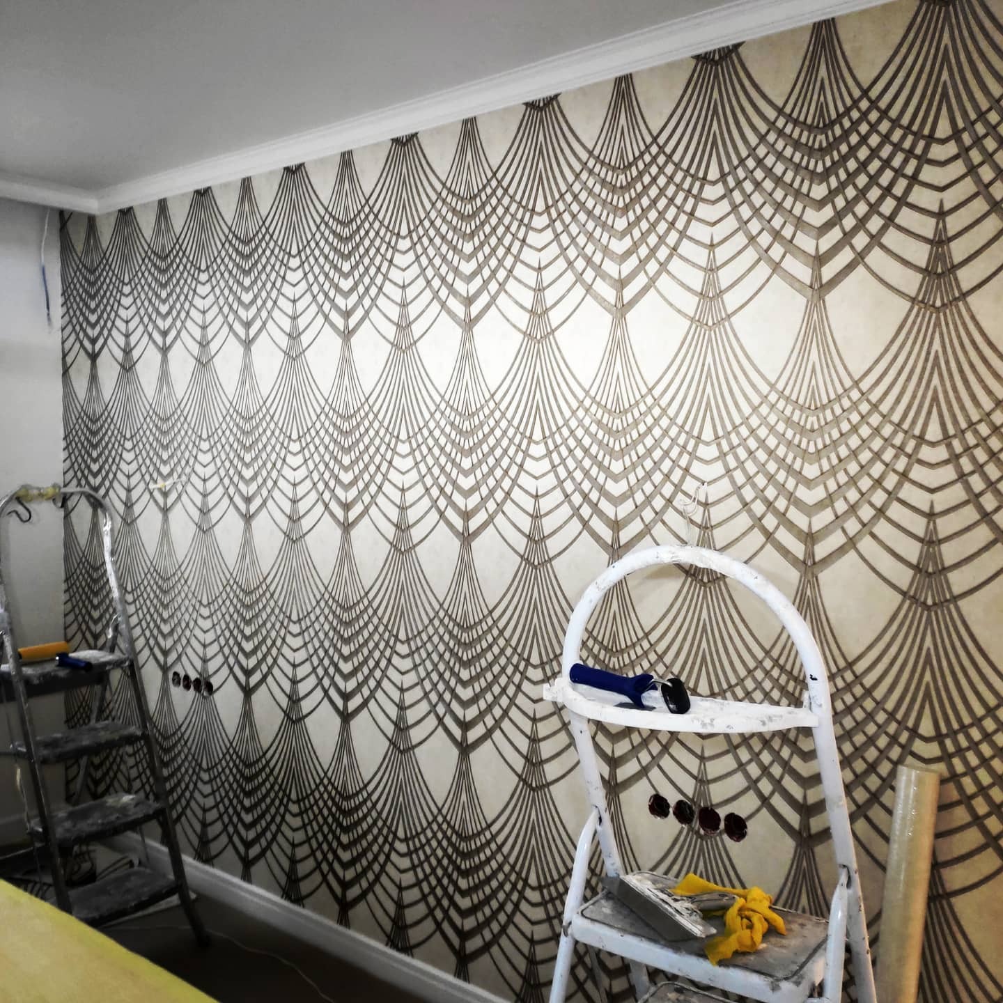 Ściana z tapetą w stylu art deco, widoczne drabiny malarskie i narzędzia. Dekoracyjne wykończenie wnętrza z wzorem wachlarzy.