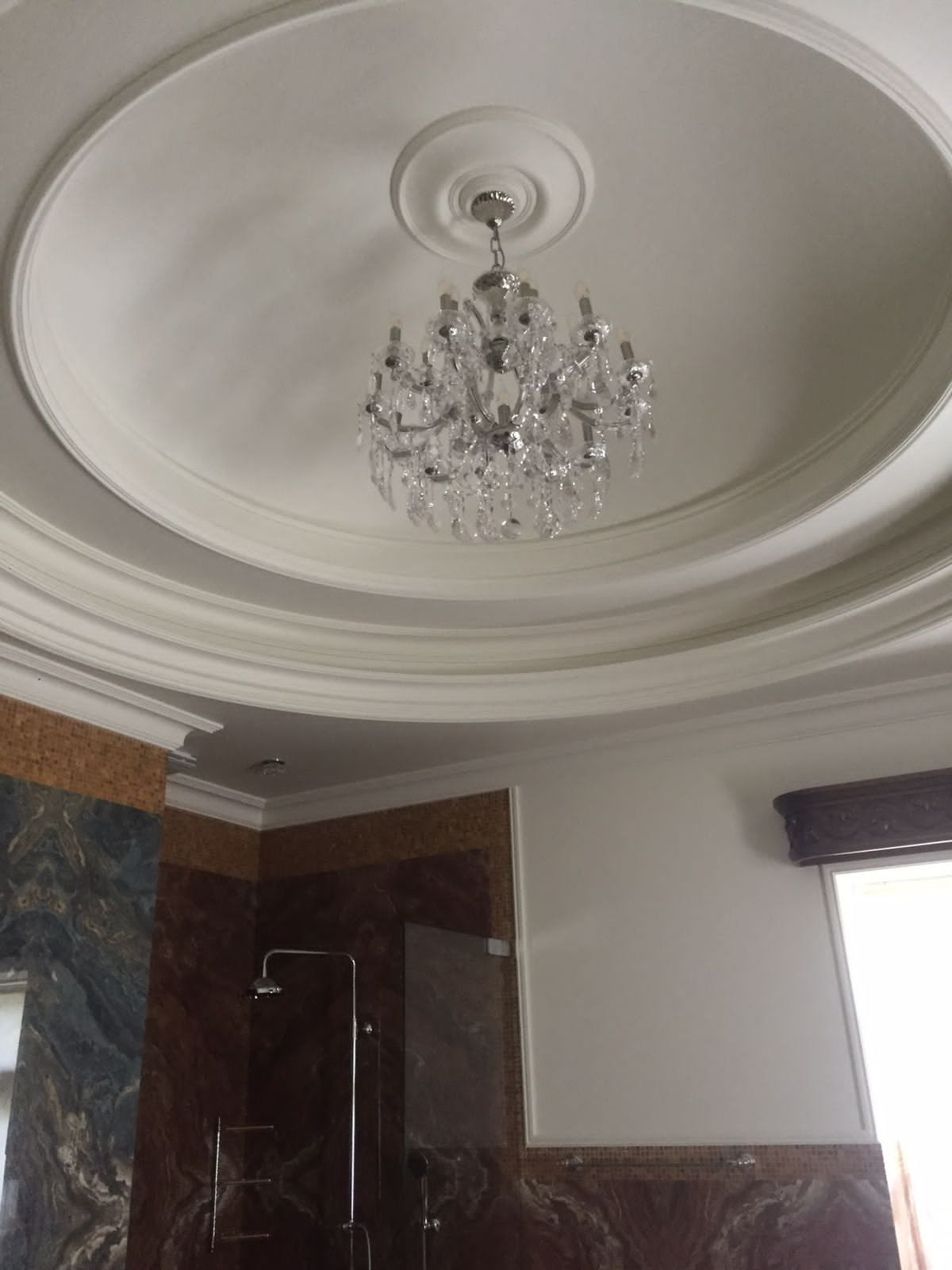 Biały sufit z dekoracyjnym gzymsem i kryształowym żyrandolem w łazience z marmurowymi ścianami i kabiną prysznicową. Eleganckie wykończenie wnętrza.