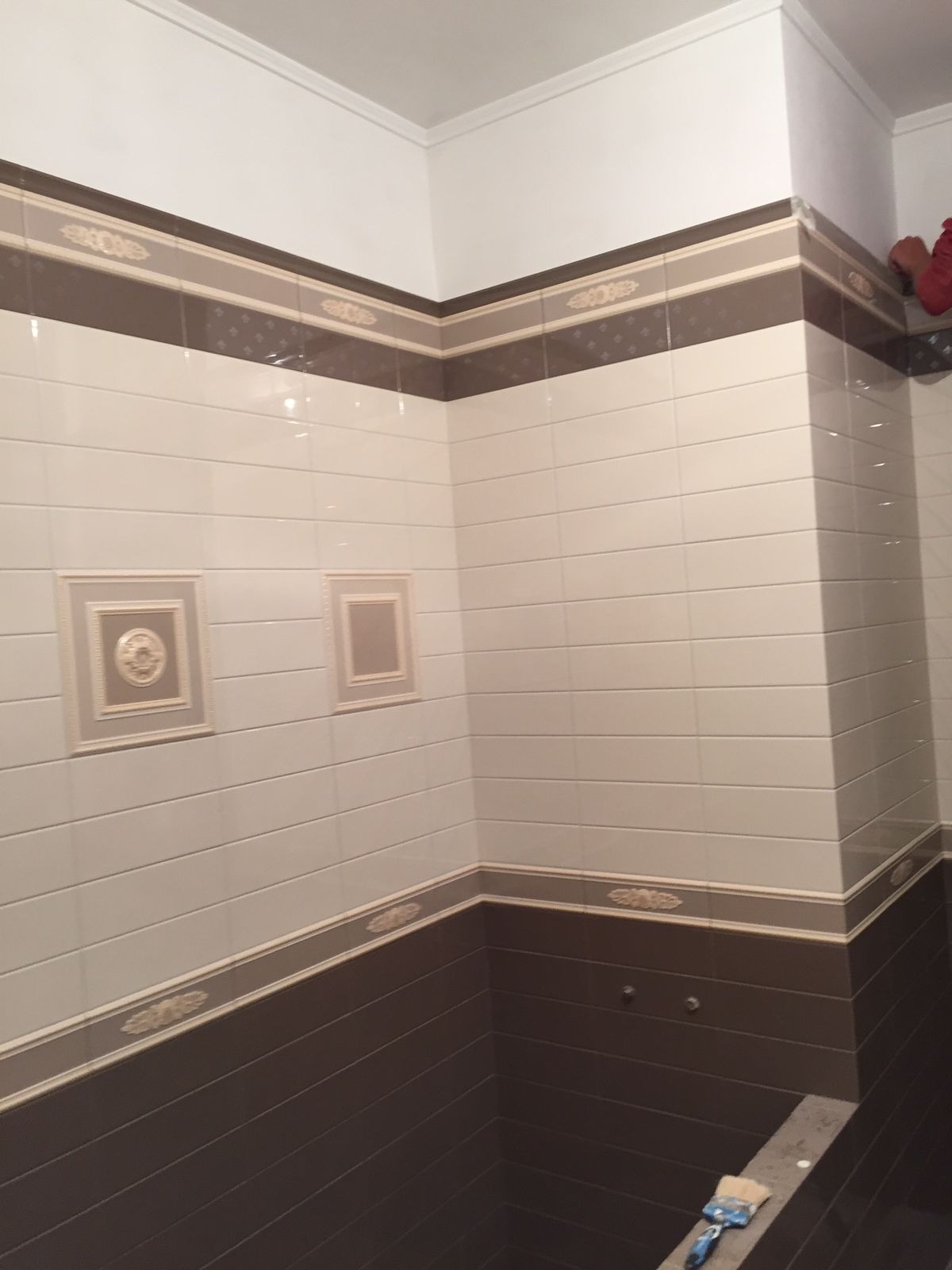 Wykończona łazienka z brązowo-białymi płytkami, dekoracyjnymi bordiurami i ozdobnymi ramkami na ścianie. Widoczny fragment sufitu z listwą. Na dole leży pędzel.