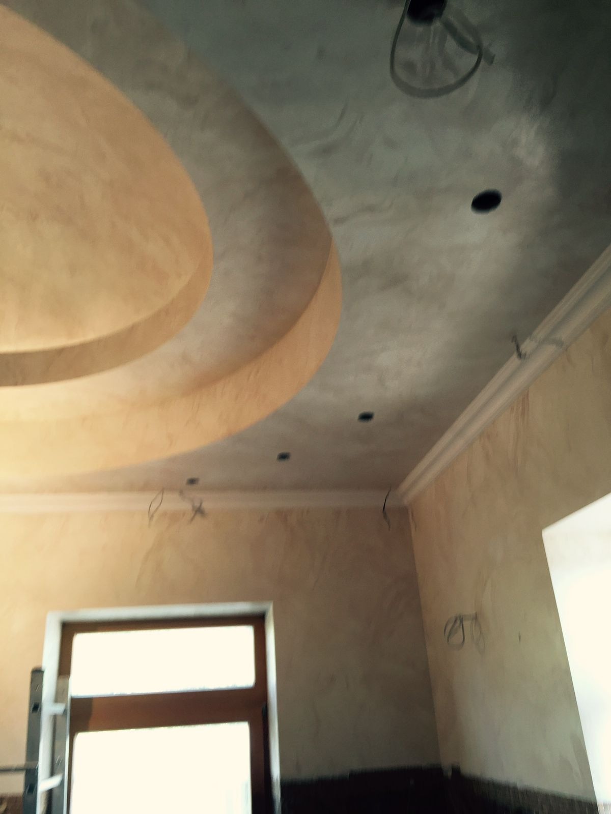 Kremowo-szary sufit z dekoracyjnym gzymsem i okrągłym wgłębieniem, widok z dołu. W tle fragment okna i ściany z fakturą. Widoczne przewody elektryczne.