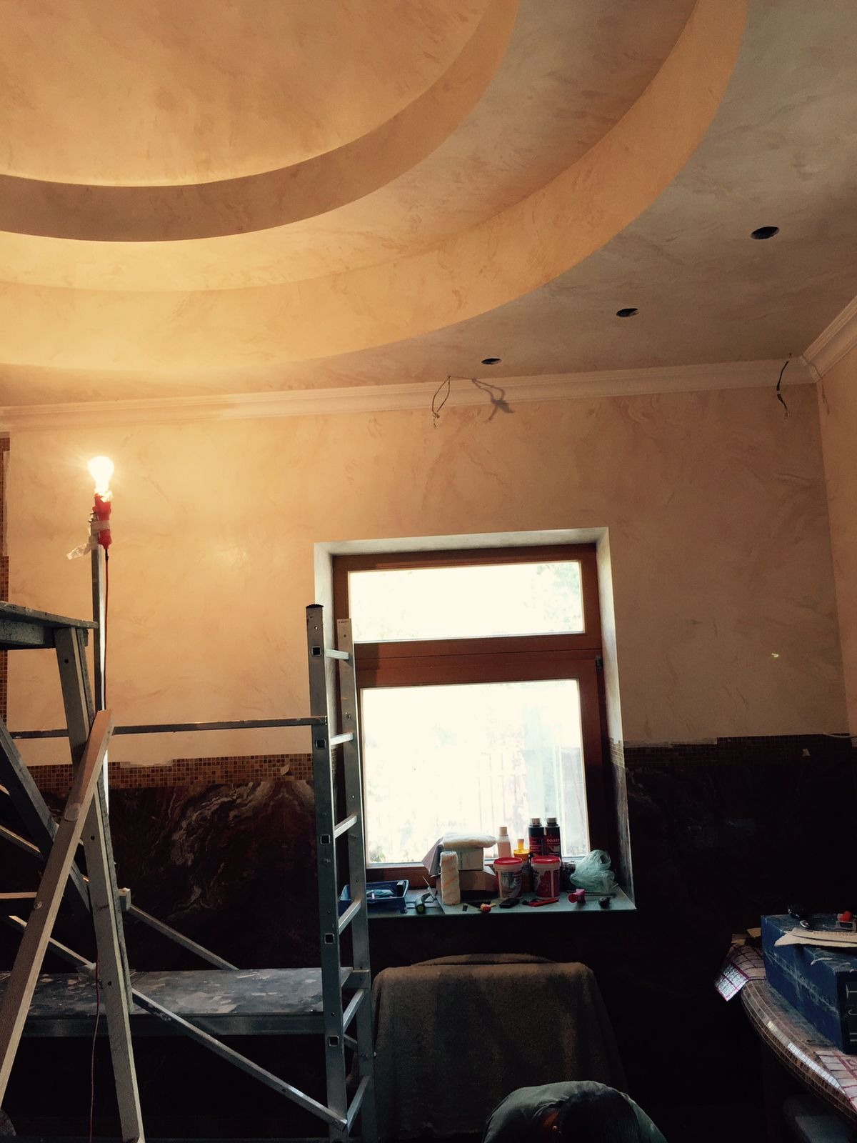 Wnętrze w trakcie prac wykończeniowych: dekoracyjny sufit z oświetleniem, ściany z efektem marmuru, drabiny i narzędzia przy oknie. Widoczne niedokończone detale.