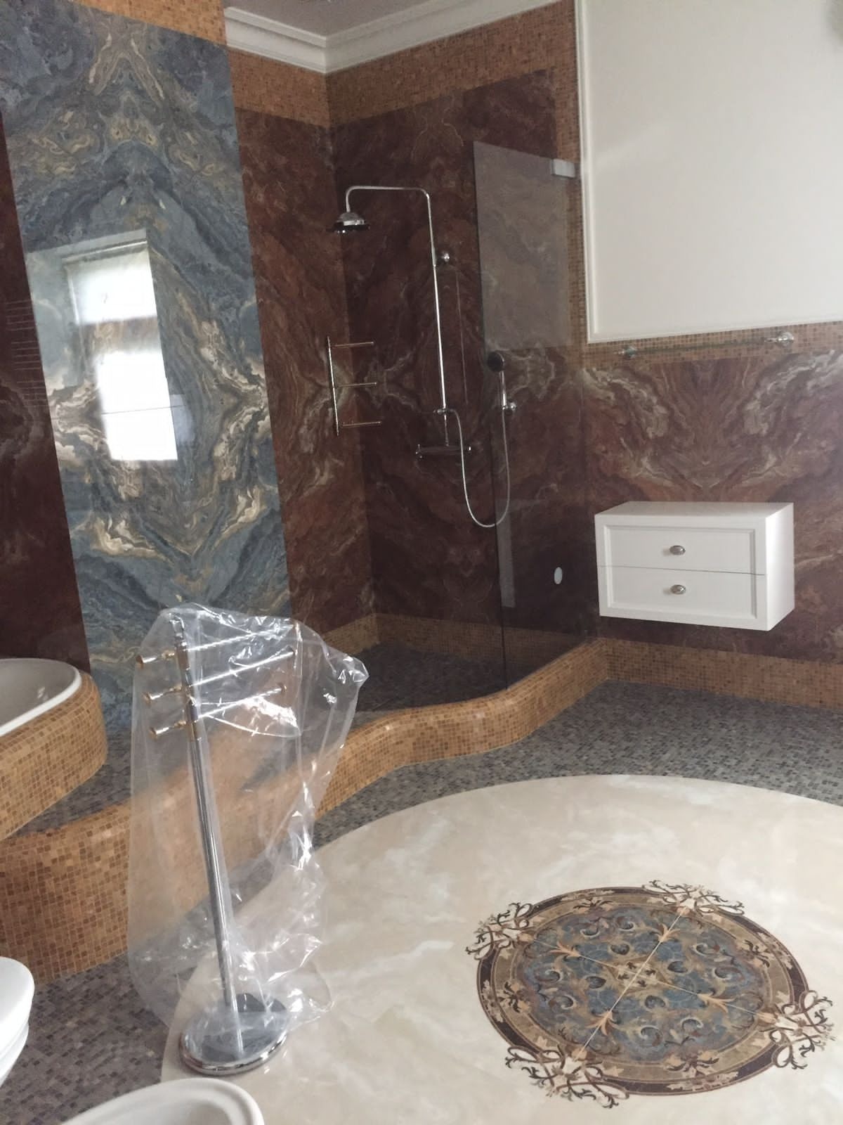 Wykończona łazienka z kabiną prysznicową typu walk-in, marmurowymi ścianami w odcieniach brązu i niebieskiego oraz okrągłą mozaiką na podłodze. Widoczna szafka wisząca.