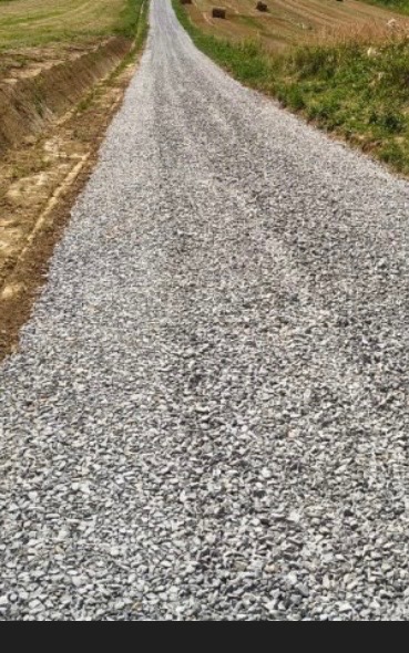 Utwardzona droga szutrowa na obszarze wiejskim, widok z perspektywy. Po bokach widoczne pola i łąki. Świeżo wysypany szuter.