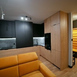 Przestrzeń z Pasją - Nowoczesna kuchnia z czarnymi i drewnianymi frontami, oświetleniem LED pod szafkami i żółtą sofą w tle. Widok na zabudowę kuchenną i część salonu.