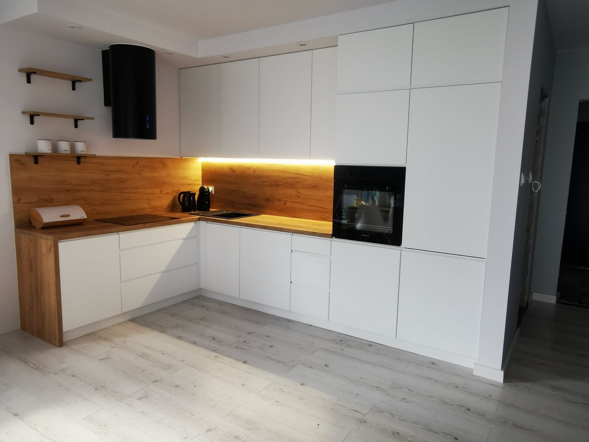 Nowoczesna kuchnia na wymiar z białymi frontami i drewnianym blatem. Oświetlenie LED pod szafkami górnymi. Czarny okap cylindryczny. Jasna podłoga imitująca drewno.