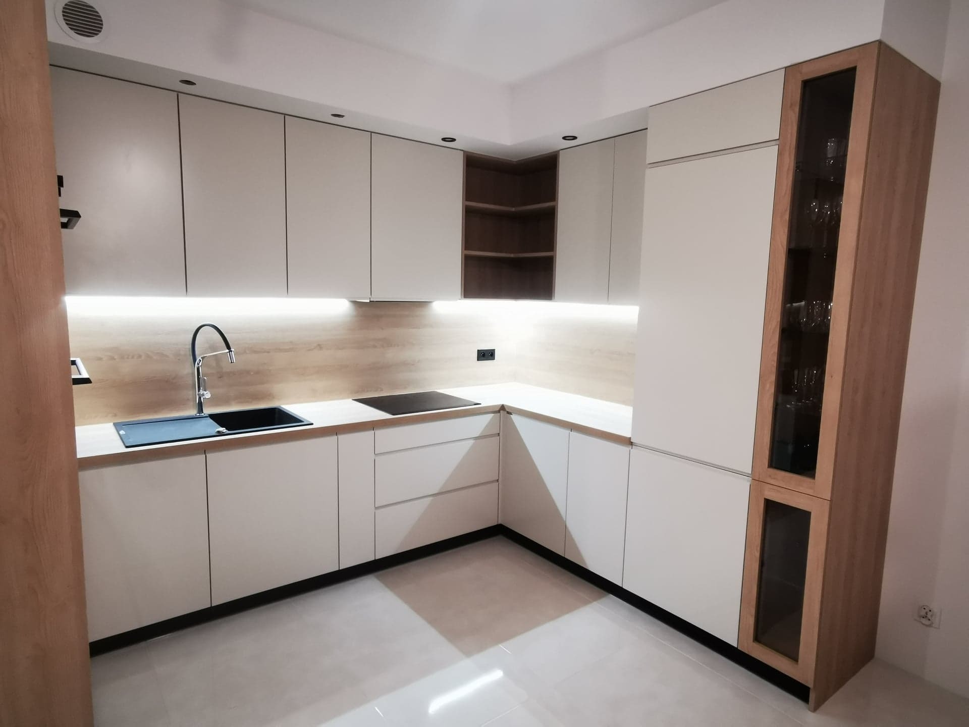 Jasna, narożna kuchnia z drewnianymi akcentami: przeszklona witryna, otwarte półki i podświetlenie LED. Minimalistyczny design z czarnym zlewem i płytą.