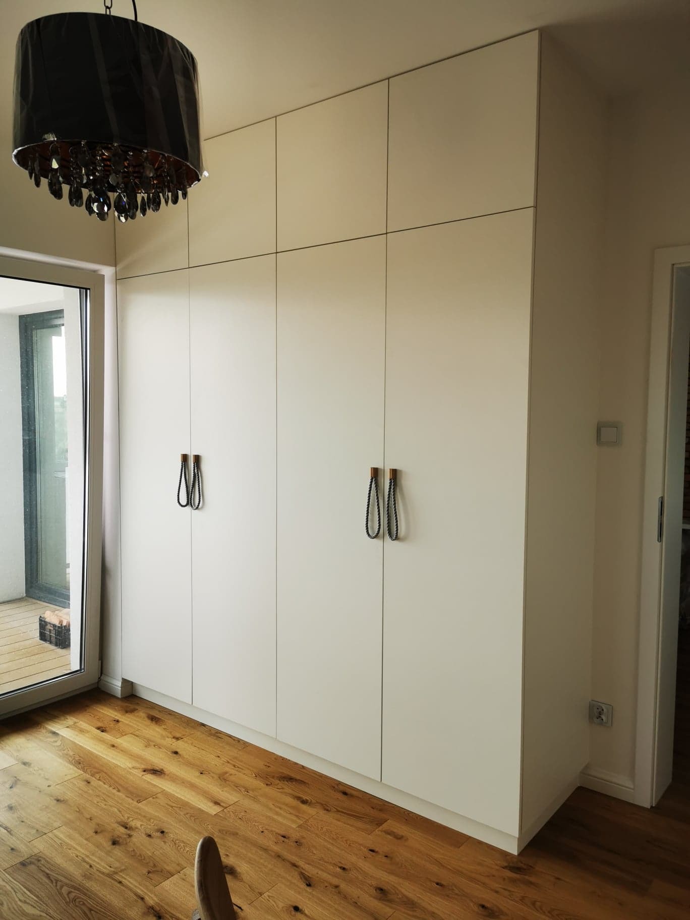 Biała szafa wnękowa z drewnianymi uchwytami sznurkowymi, parkiet, widok na balkon przez okno. Minimalistyczny design, oświetlenie lampą wiszącą.
