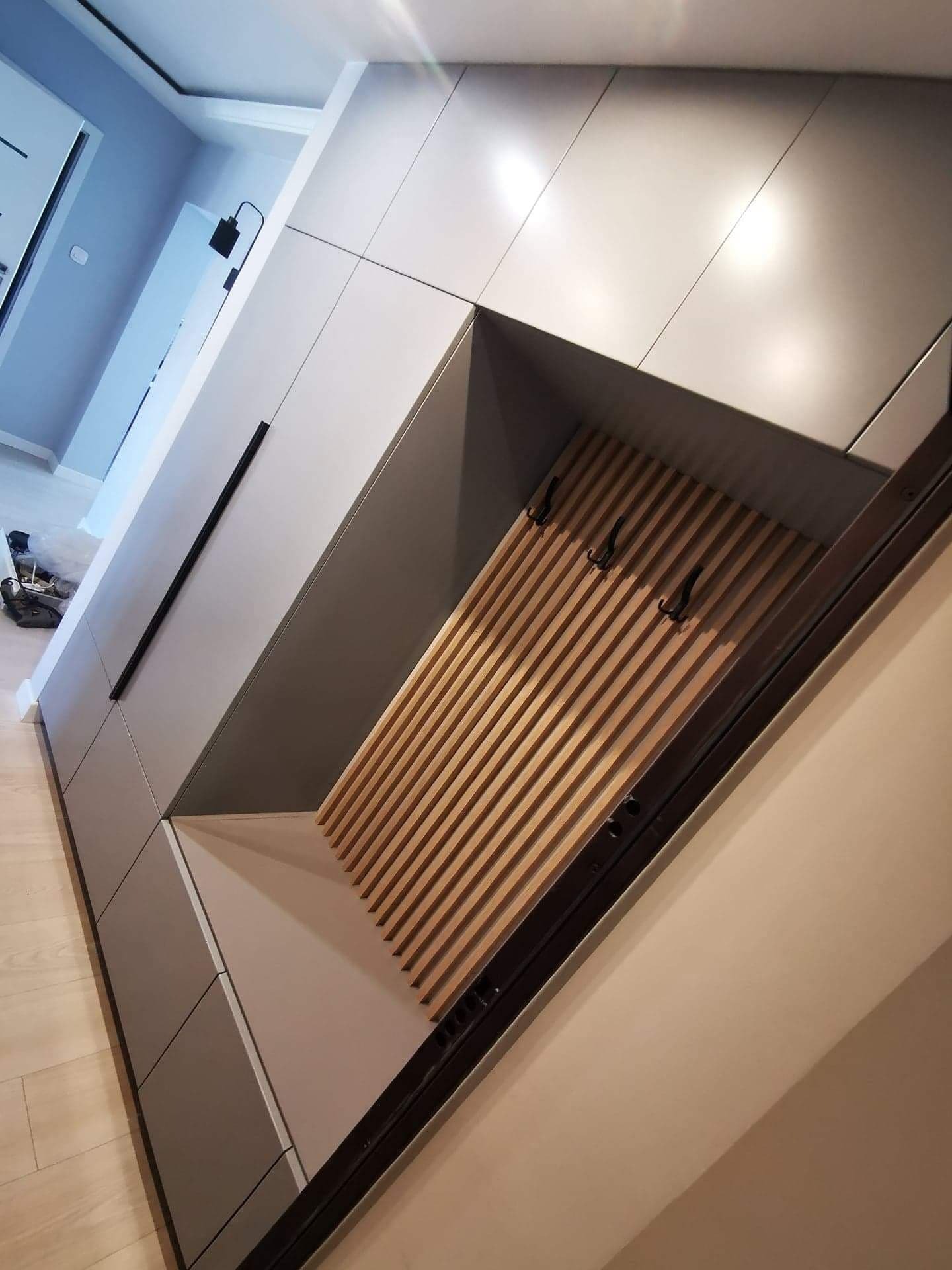 Szara szafa wnękowa z drewnianymi lamelami i czarnymi haczykami na ubrania, wkomponowana w przedpokój. Minimalistyczny design, nowoczesne wykończenie.