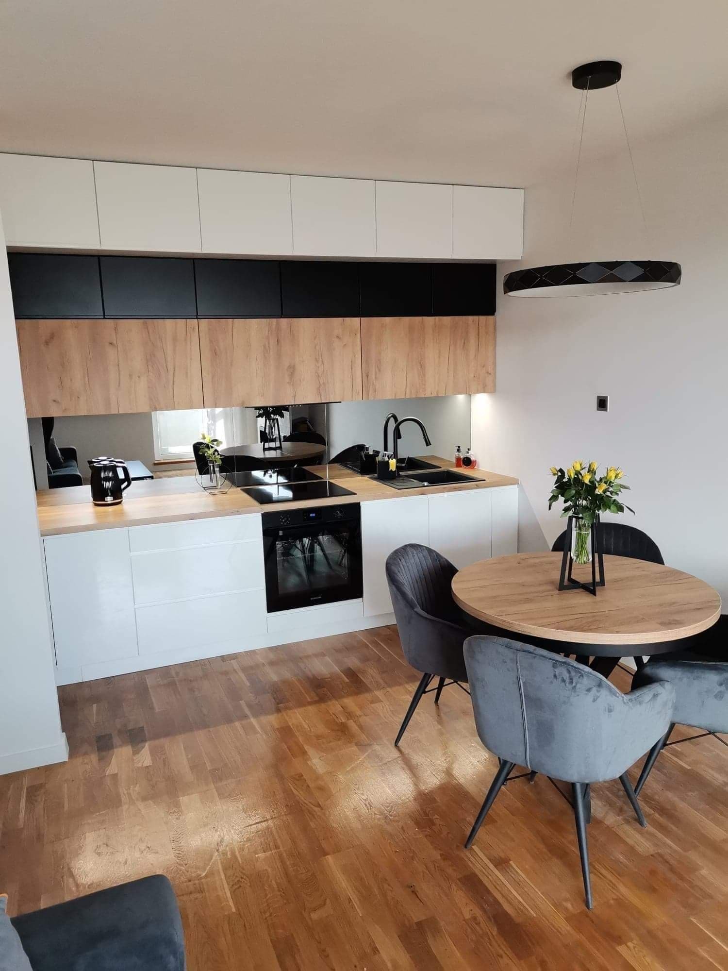 Nowoczesna kuchnia z lustrzanym panelem nad blatem, drewnianymi szafkami i okrągłym stołem z szarymi krzesłami. Minimalistyczny design, ciepłe oświetlenie.