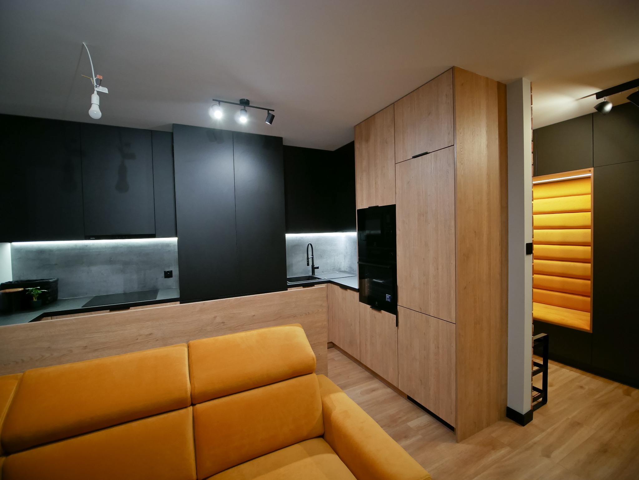 Nowoczesna kuchnia z czarnymi i drewnianymi frontami, oświetleniem LED pod szafkami i żółtą sofą w tle. Widok na zabudowę kuchenną i część salonu.