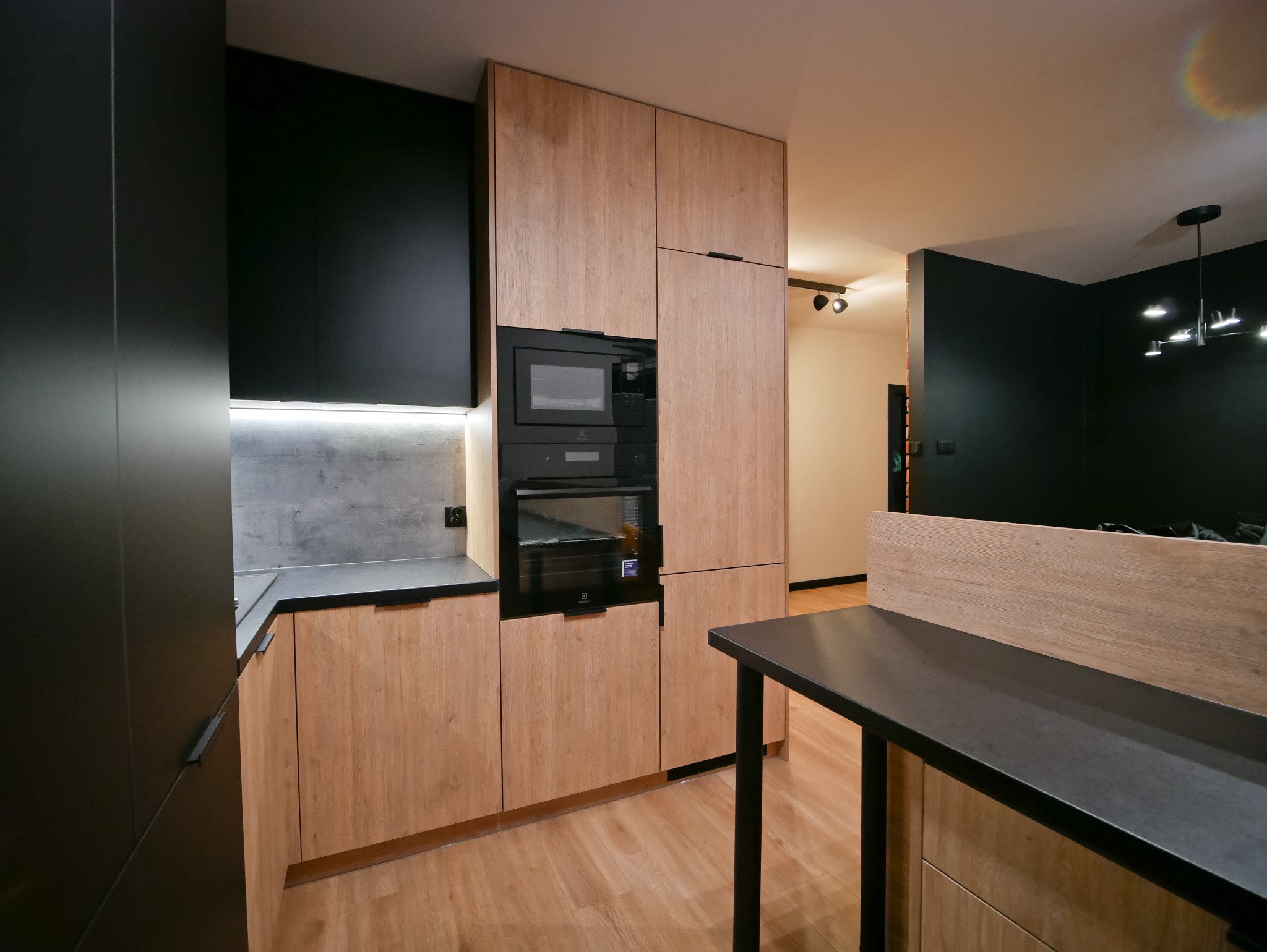 Nowoczesna kuchnia z drewnianymi frontami i czarnymi akcentami. Zabudowa kuchenna z piekarnikiem i mikrofalówką. Oświetlenie LED pod szafkami. Minimalistyczny design.