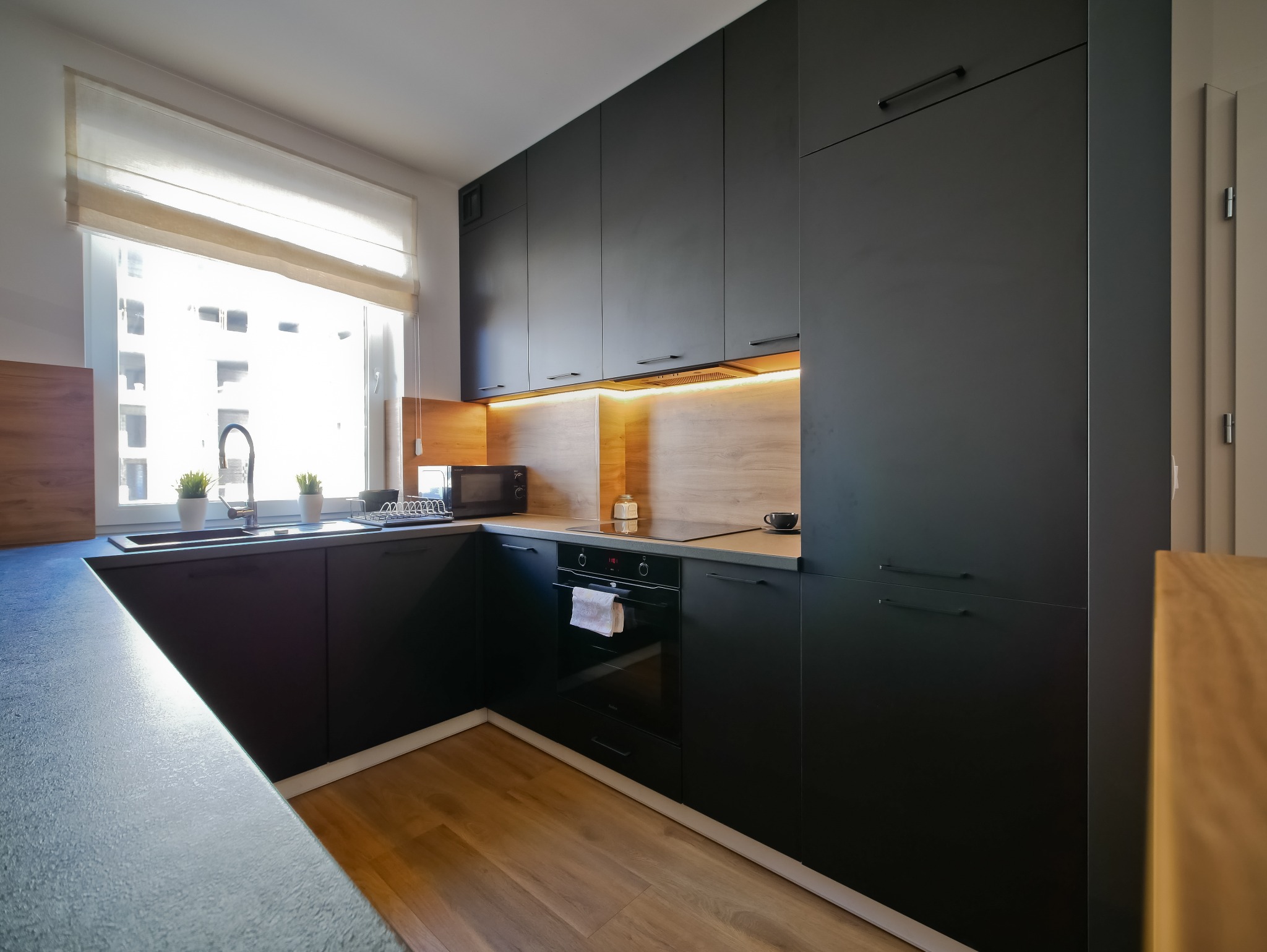 Nowoczesna kuchnia z czarnymi matowymi frontami, drewnianym panelem i oświetleniem LED. Minimalistyczny design, jasne wnętrze z oknem i roślinami.