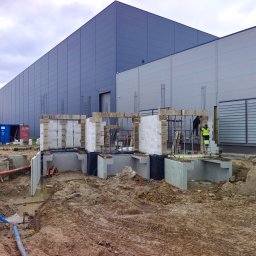 BKR Inżynieria Bartosz Krych - Budowa hali: fundamenty zbrojone, ściany z bloczków, pracownik w kamizelce na drabinie. Widoczne wykopy, rury i kontenery. Duży budynek w tle, pochmurne niebo.