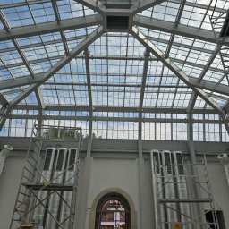 BKR Inżynieria Bartosz Krych - Szklany dach z metalową konstrukcją w budynku w trakcie prac wykończeniowych. Widoczne rusztowania i instalacje wentylacyjne. Osoba w kamizelce odblaskowej w tle.