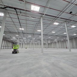 BKR Inżynieria Bartosz Krych - Przestronne wnętrze hali magazynowej z betonową posadzką, białymi kolumnami i dachem z licznymi świetlikami. Widoczny podnośnik nożycowy i pracownik w oddali.