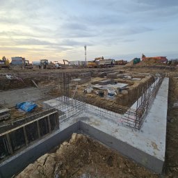 BKR Inżynieria - Fundamenty domu jednorodzinnego w trakcie budowy. Widoczne zbrojenia, szalunki i wykop. W tle maszyny budowlane i pochmurne niebo o zachodzie słońca.