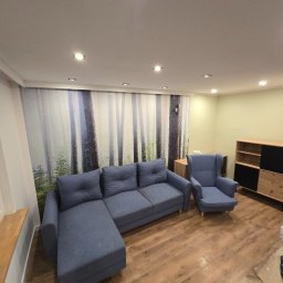 BKR Inżynieria Bartosz Krych - Jasne wnętrze po remoncie: narożnik z fotelami na drewnianej podłodze, ściana z fototapetą leśną, nowoczesna komoda. Widoczne ślady prac wykończeniowych.