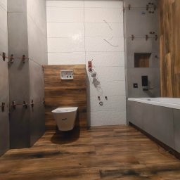 BKR Inżynieria Bartosz Krych - Wnętrze łazienki w trakcie wykańczania: szare i białe płytki na ścianach, drewnopodobna podłoga, wisząca toaleta. Widoczne krzyżyki dystansowe na płytkach.