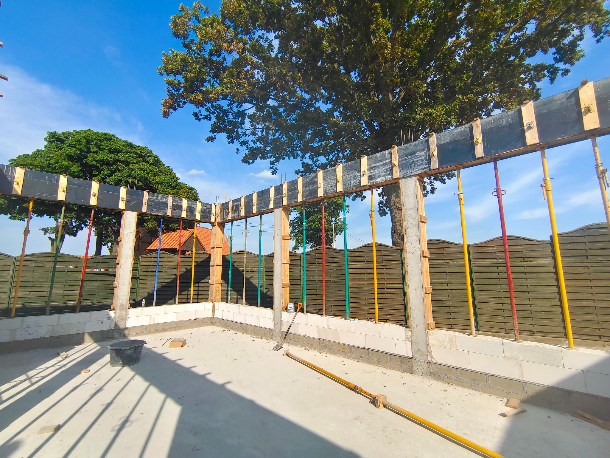 Fundamenty domu w trakcie budowy, z szalunkiem i podporami. Widoczne bloczki fundamentowe, betonowa wylewka i narzędzia budowlane. W tle drzewa i ogrodzenie.