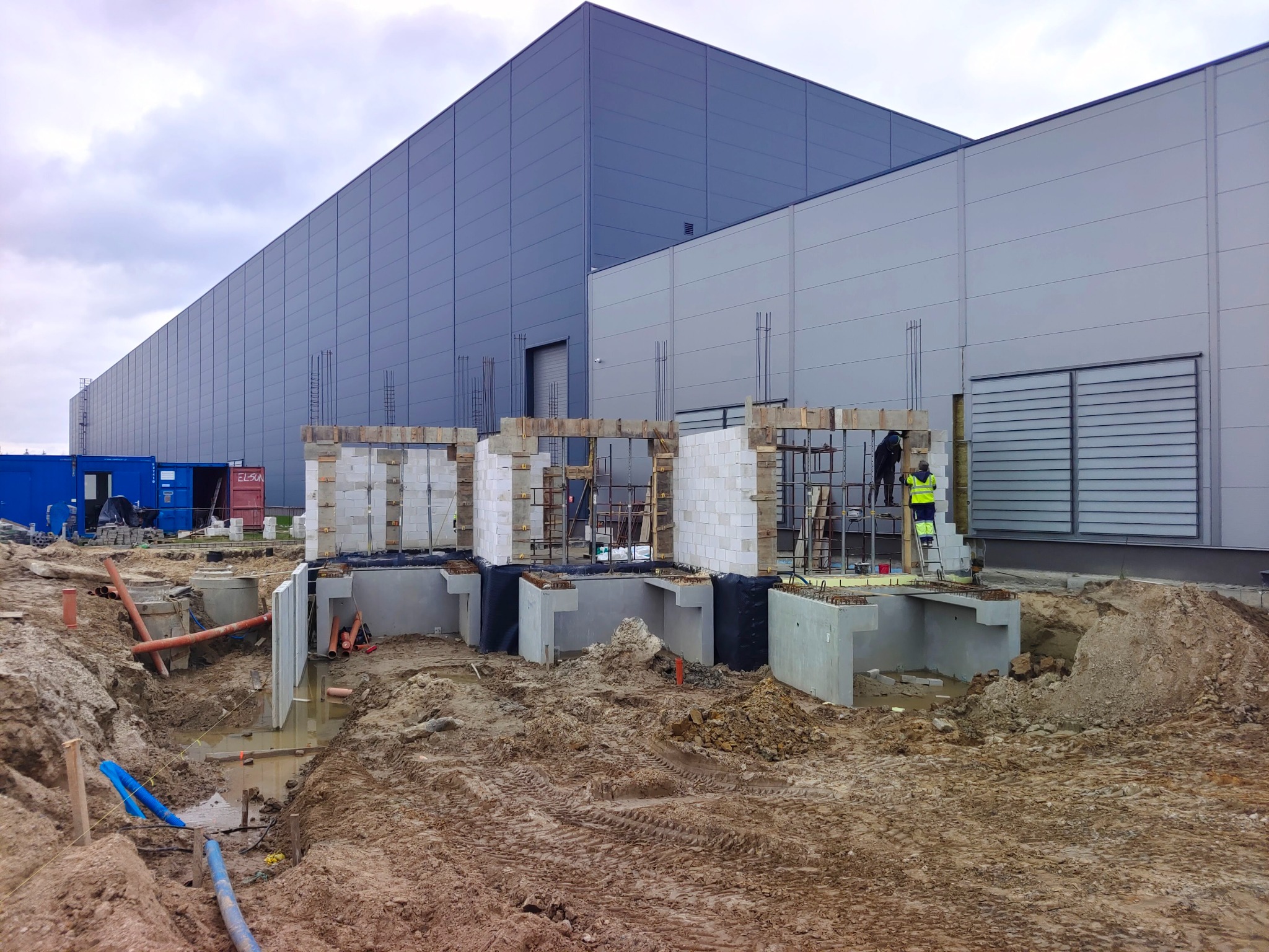 Budowa hali: fundamenty zbrojone, ściany z bloczków, pracownik w kamizelce na drabinie. Widoczne wykopy, rury i kontenery. Duży budynek w tle, pochmurne niebo.