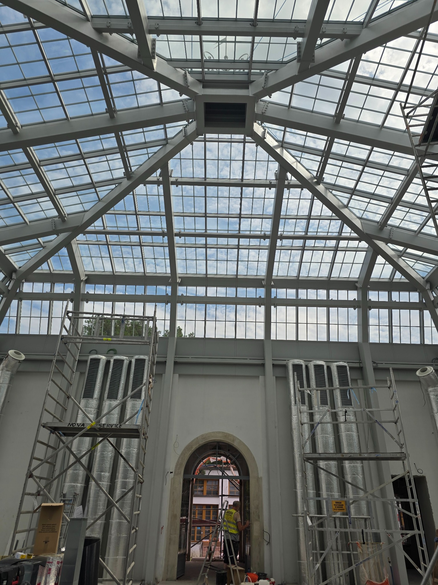 Szklany dach z metalową konstrukcją w budynku w trakcie prac wykończeniowych. Widoczne rusztowania i instalacje wentylacyjne. Osoba w kamizelce odblaskowej w tle.