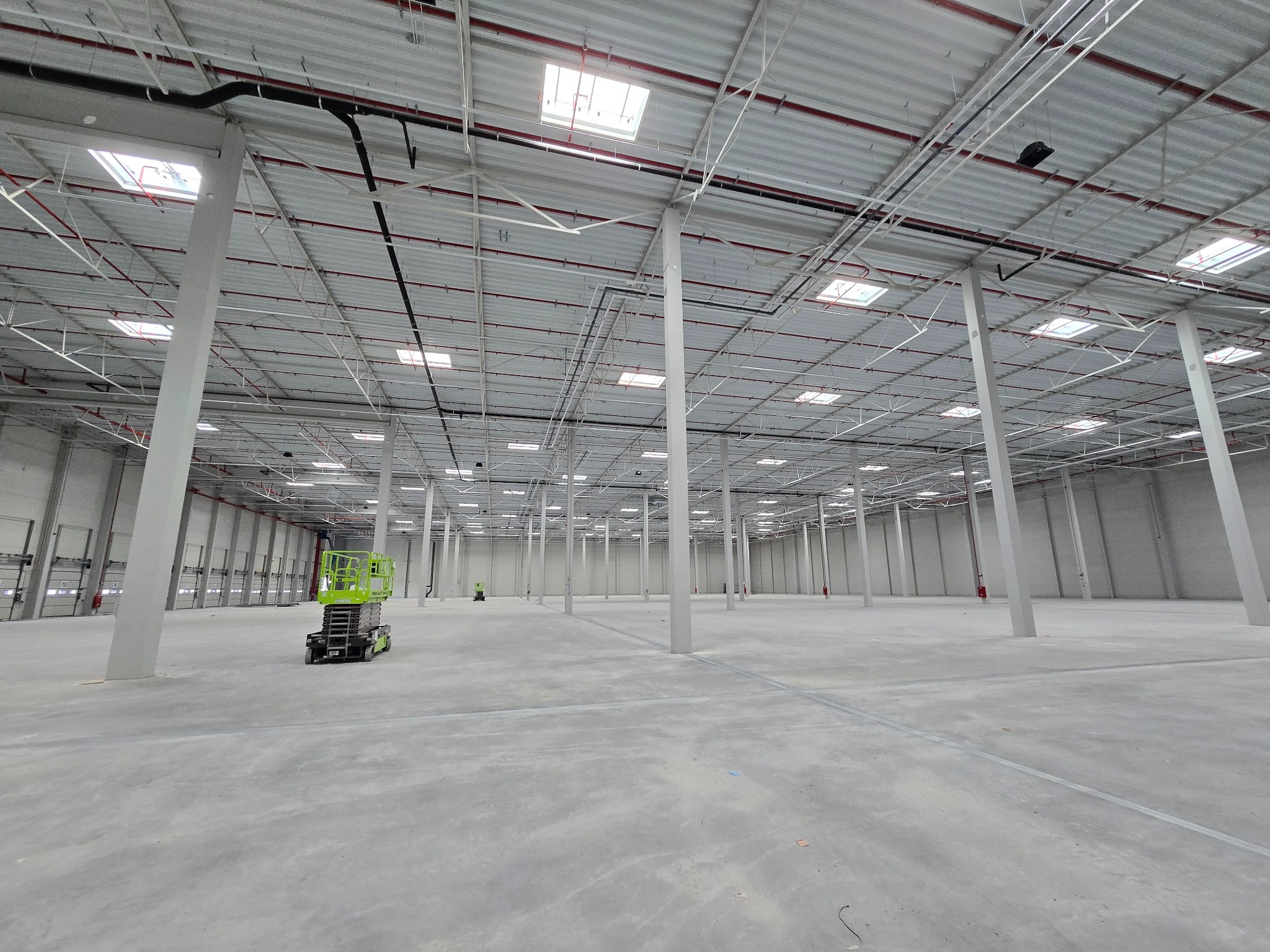 Przestronne wnętrze hali magazynowej z betonową posadzką, białymi kolumnami i dachem z licznymi świetlikami. Widoczny podnośnik nożycowy i pracownik w oddali.