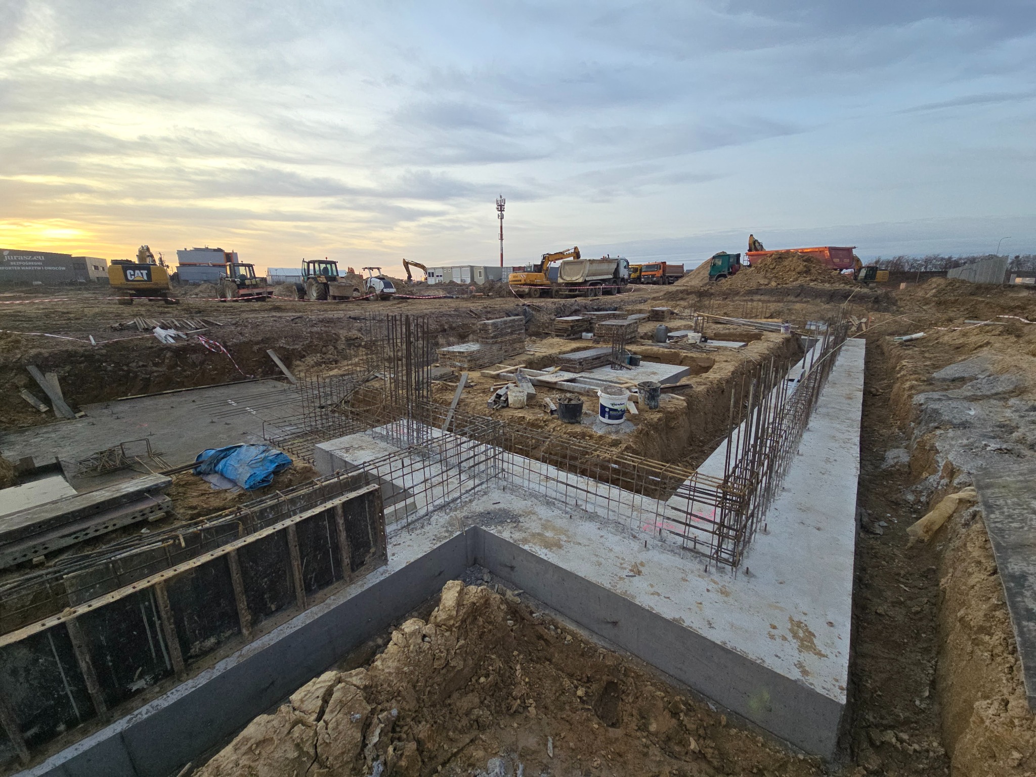 Fundamenty domu jednorodzinnego w trakcie budowy. Widoczne zbrojenia, szalunki i wykop. W tle maszyny budowlane i pochmurne niebo o zachodzie słońca.