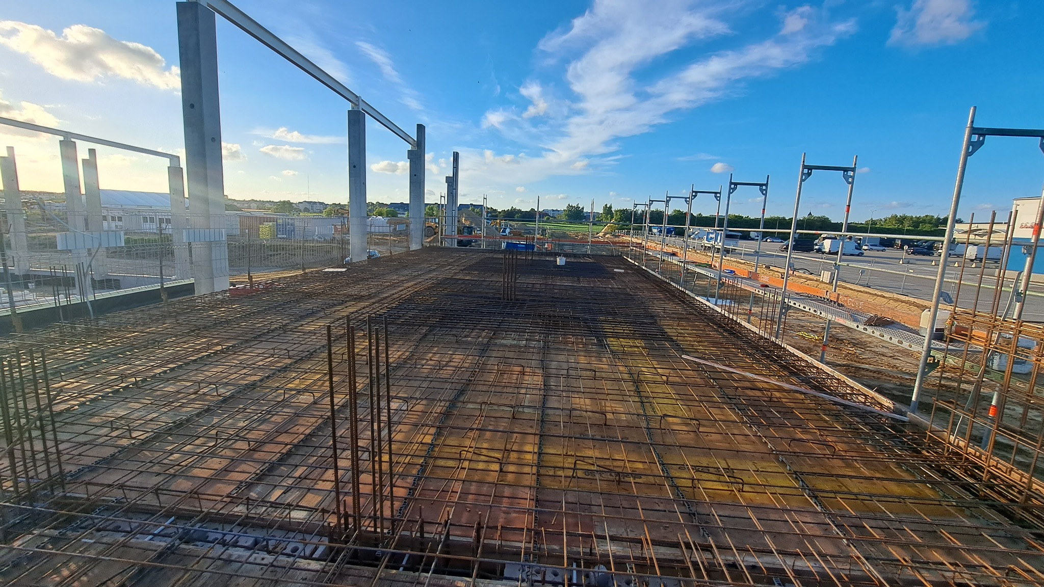 Zbrojenie płyty fundamentowej na budowie hali, widoczne stalowe słupy konstrukcyjne i rusztowania. Jasne niebo w tle.