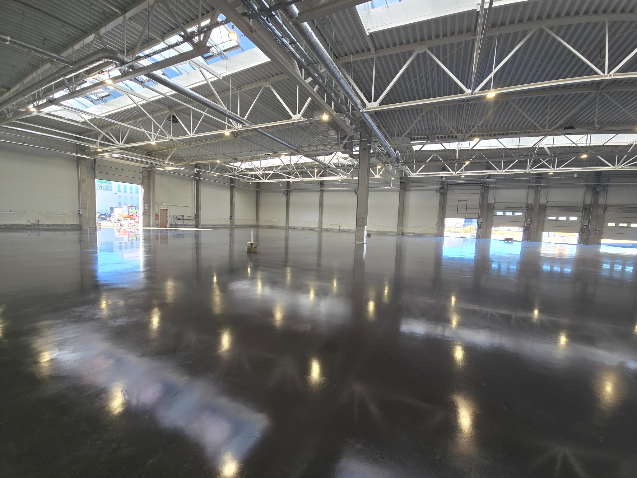 Polerowana, szara posadzka przemysłowa w hali z metalową konstrukcją dachu i świetlikami. Widoczne bramy wjazdowe i betonowe ściany.