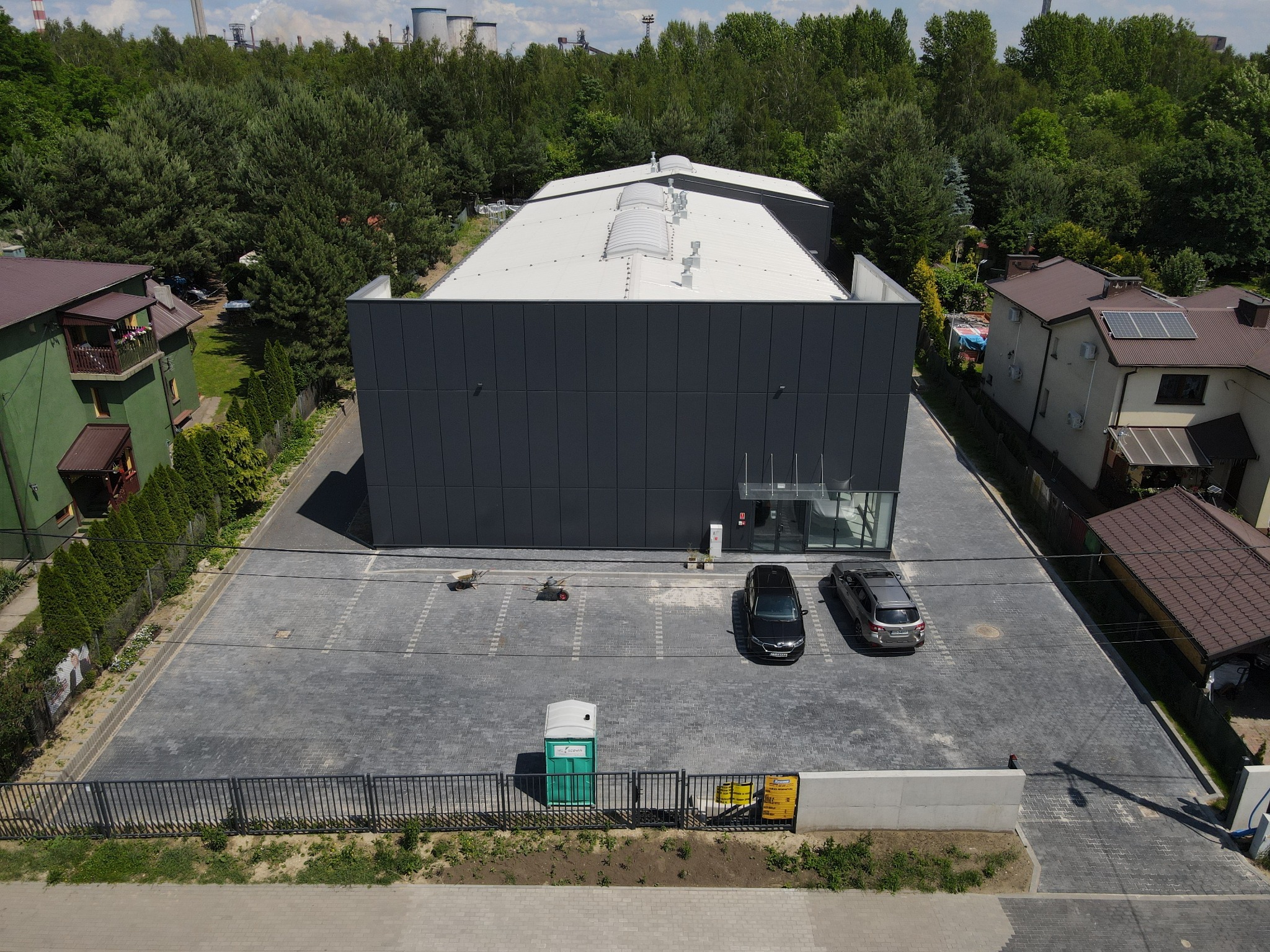 Nowoczesny budynek biurowy z szarą elewacją i białym dachem, widziany z góry. Na parkingu dwa samochody i przenośna toaleta. W tle drzewa i kominy przemysłowe.