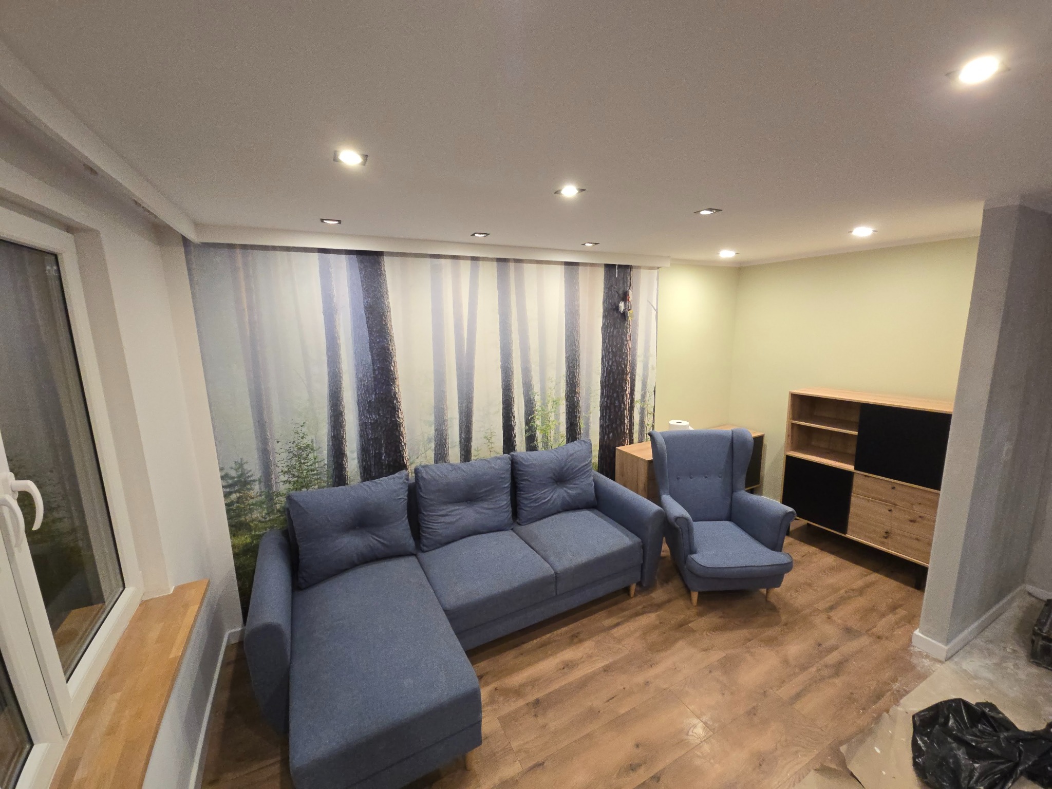 Jasne wnętrze po remoncie: narożnik z fotelami na drewnianej podłodze, ściana z fototapetą leśną, nowoczesna komoda. Widoczne ślady prac wykończeniowych.