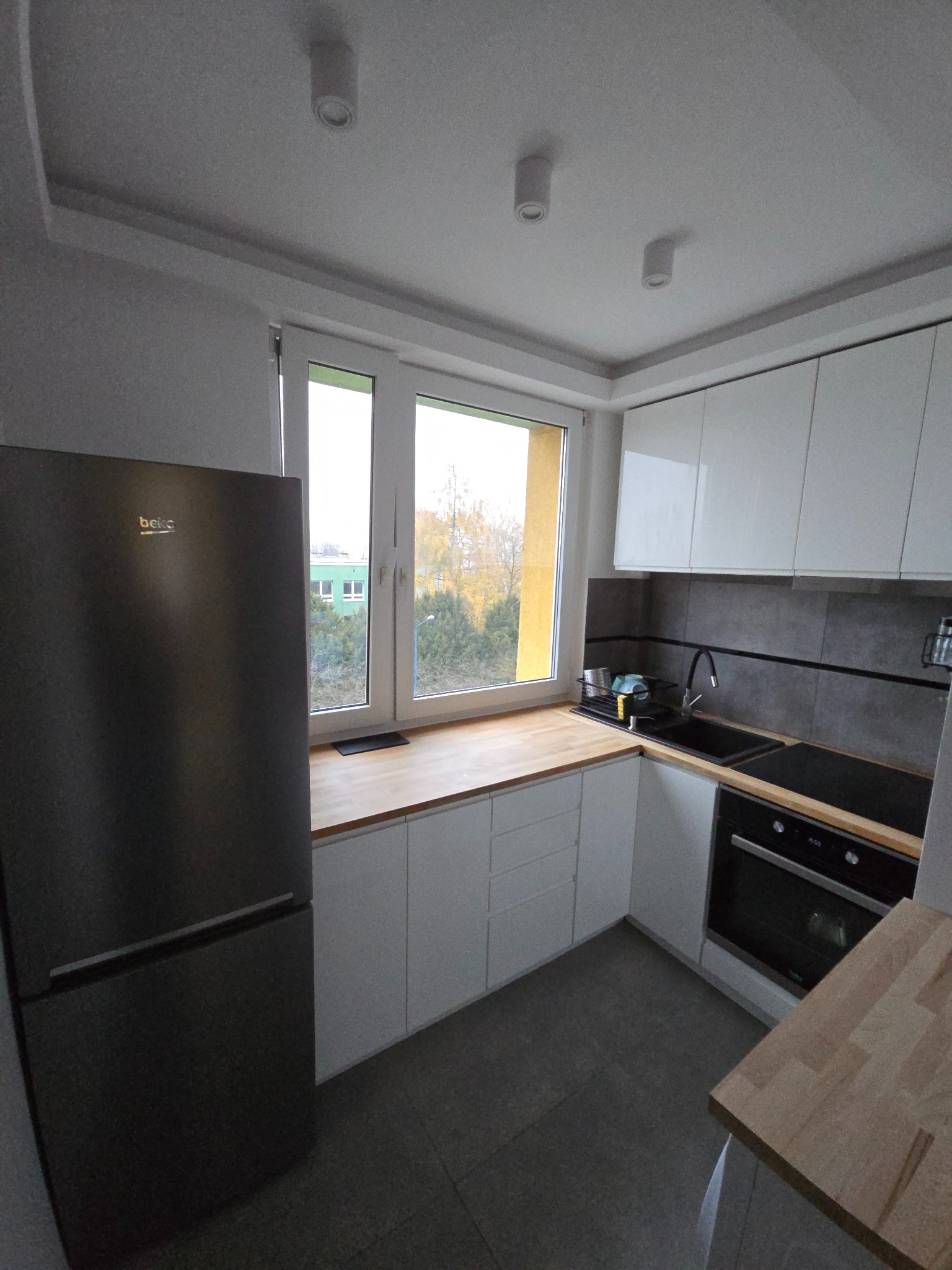 Nowoczesna kuchnia z białymi szafkami, drewnianym blatem i czarną lodówką Beko. Widok na jesienne drzewa za oknem. Minimalistyczny design i funkcjonalne rozwiązania.