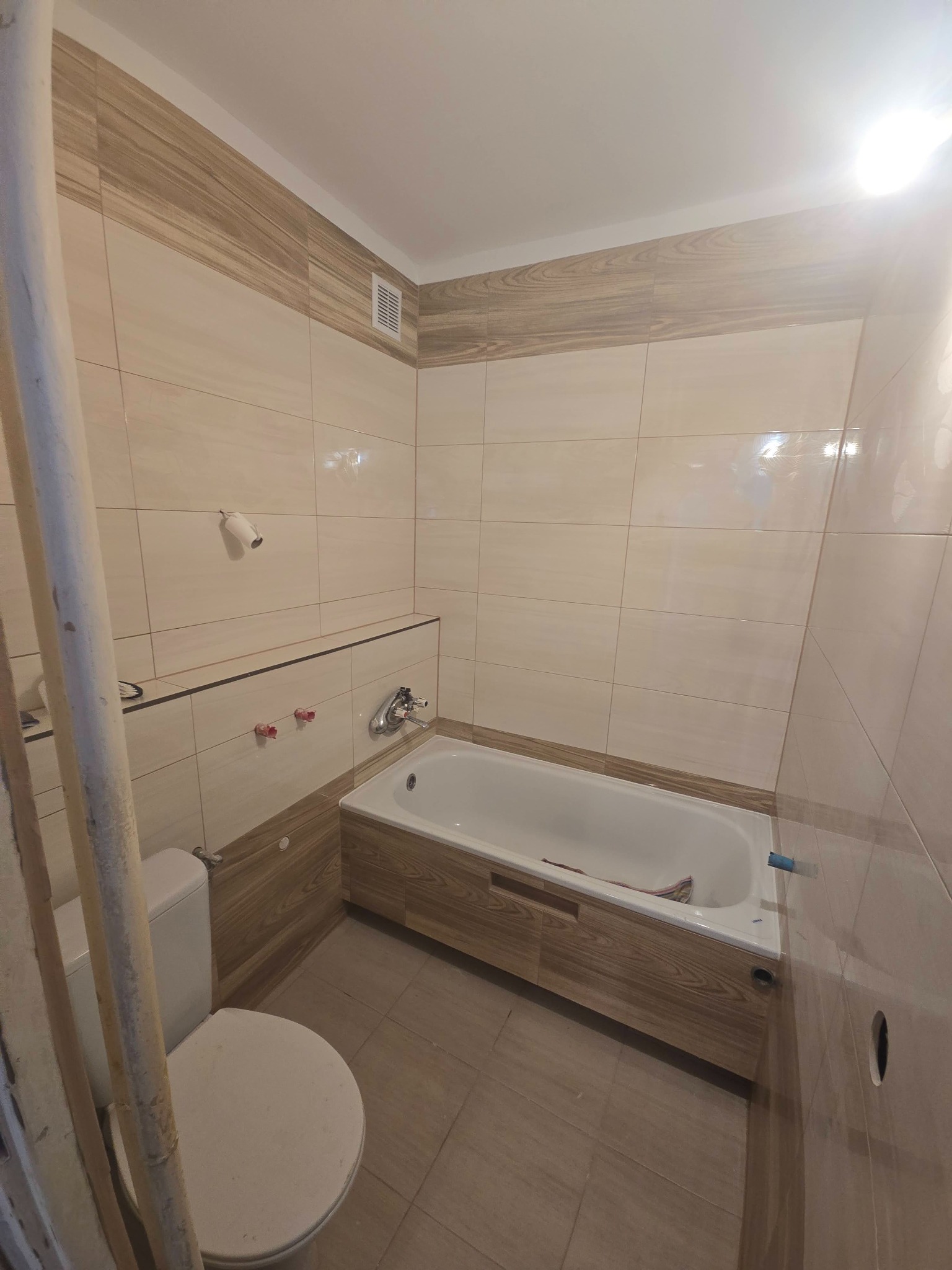 Jasna łazienka po remoncie: beżowe płytki z drewnopodobnym pasem, biała wanna z obudową imitującą drewno, toaleta. Widoczne rury i niedokończone detale.