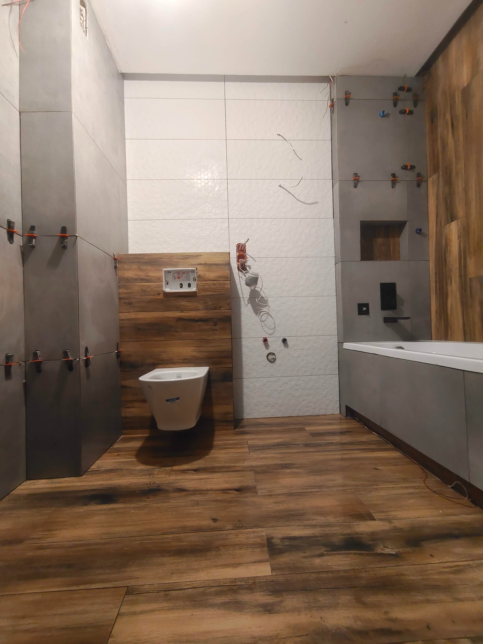 Wnętrze łazienki w trakcie wykańczania: szare i białe płytki na ścianach, drewnopodobna podłoga, wisząca toaleta. Widoczne krzyżyki dystansowe na płytkach.