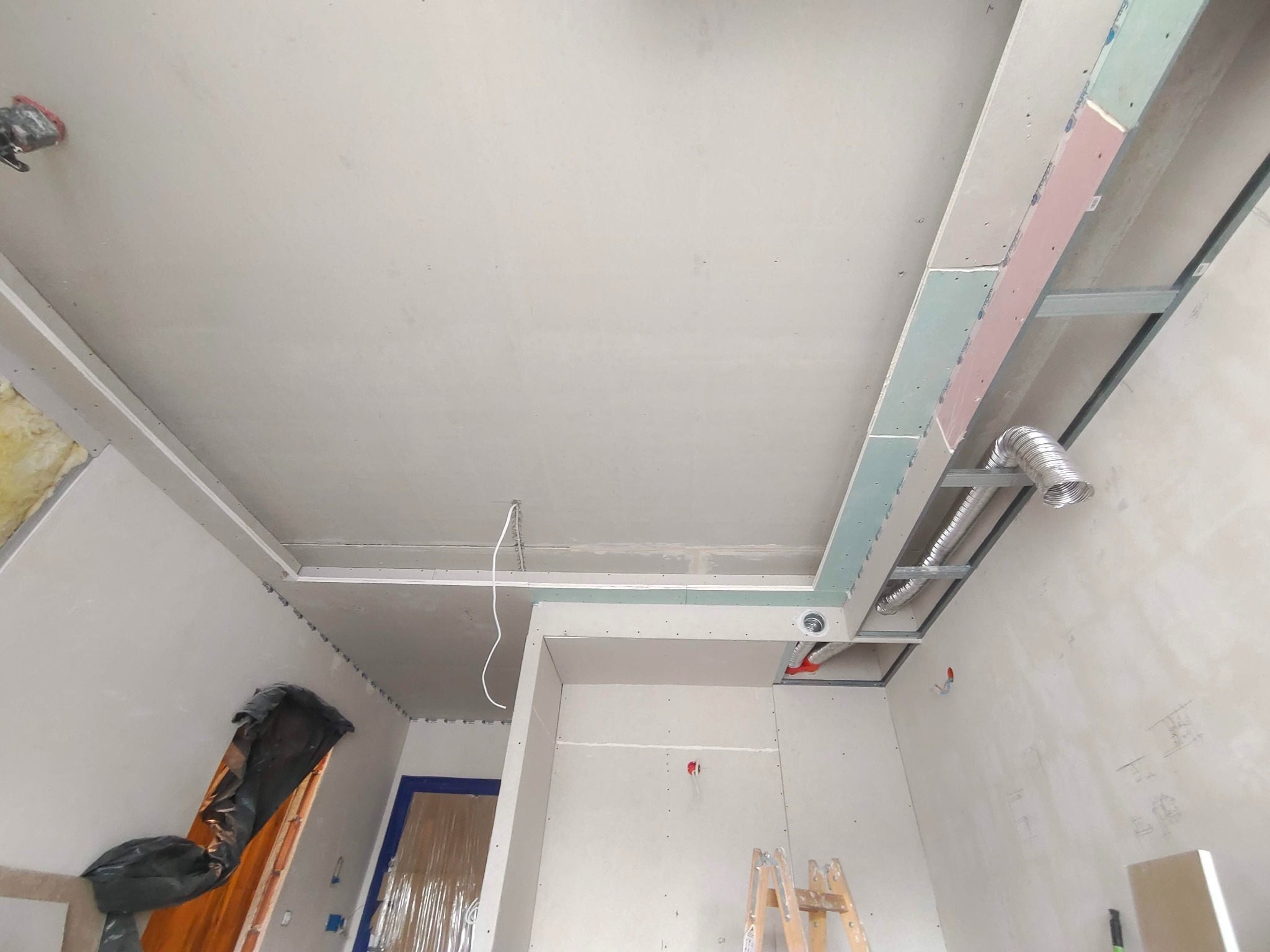 Sufit podwieszany z widoczną konstrukcją metalową i fragmentami płyt gipsowo-kartonowych. Widoczne elementy wentylacji i instalacji elektrycznej. Ściany w trakcie szpachlowania.