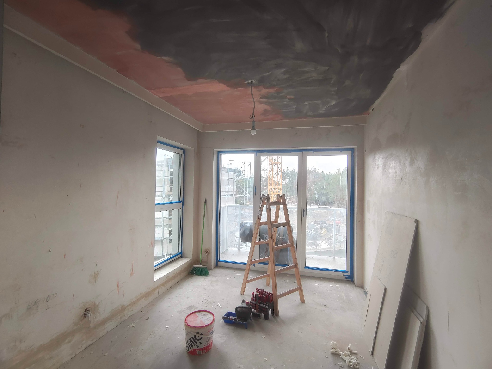 Pokój w trakcie malowania: ściany przygotowane, sufit częściowo pomalowany na szaro, drabina, pędzle, wałki i wiadro farby na podłodze. Widok z okna na budowę.