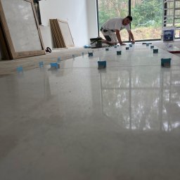 FachNaTak - Glazurnik układa jasne płytki podłogowe, używając systemu poziomowania. Widoczne narzędzia i duże okno z widokiem na zieleń. Praca w toku, wnętrze w stanie surowym.