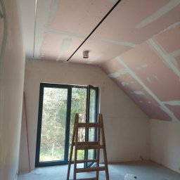 FachNaTak - Poddasze w trakcie prac: płyty karton-gips na skosach, częściowo szpachlowane łączenia, drabina, okno z widokiem na zieleń, farby i wałki malarskie na podłodze.