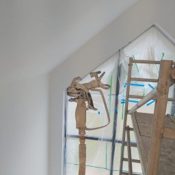 FachNaTak - Malowanie natryskowe ścian w pokoju ze skosami. Widoczny pistolet natryskowy, drabina i okno zabezpieczone folią. Ściany świeżo pomalowane na biało.
