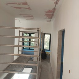 FachNaTak - Wnętrze w trakcie prac wykończeniowych: szpachlowana sufit z widocznymi łatami, metalowe rusztowanie, przygotowane do malowania ściany i futryny drzwiowe.