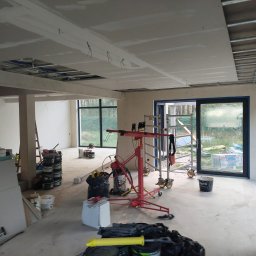 FachNaTak - Montaż płyt kartonowo-gipsowych na suficie w przestronnym wnętrzu. Widoczny podnośnik do płyt, metalowa konstrukcja sufitu i częściowo wykończone ściany.