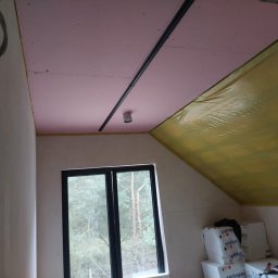 FachNaTak - Poddasze z oknem: różowa płyta karton-gips na suficie, czarna szyna oświetleniowa, żółta folia izolacyjna na skosie. Widoczne materiały budowlane.