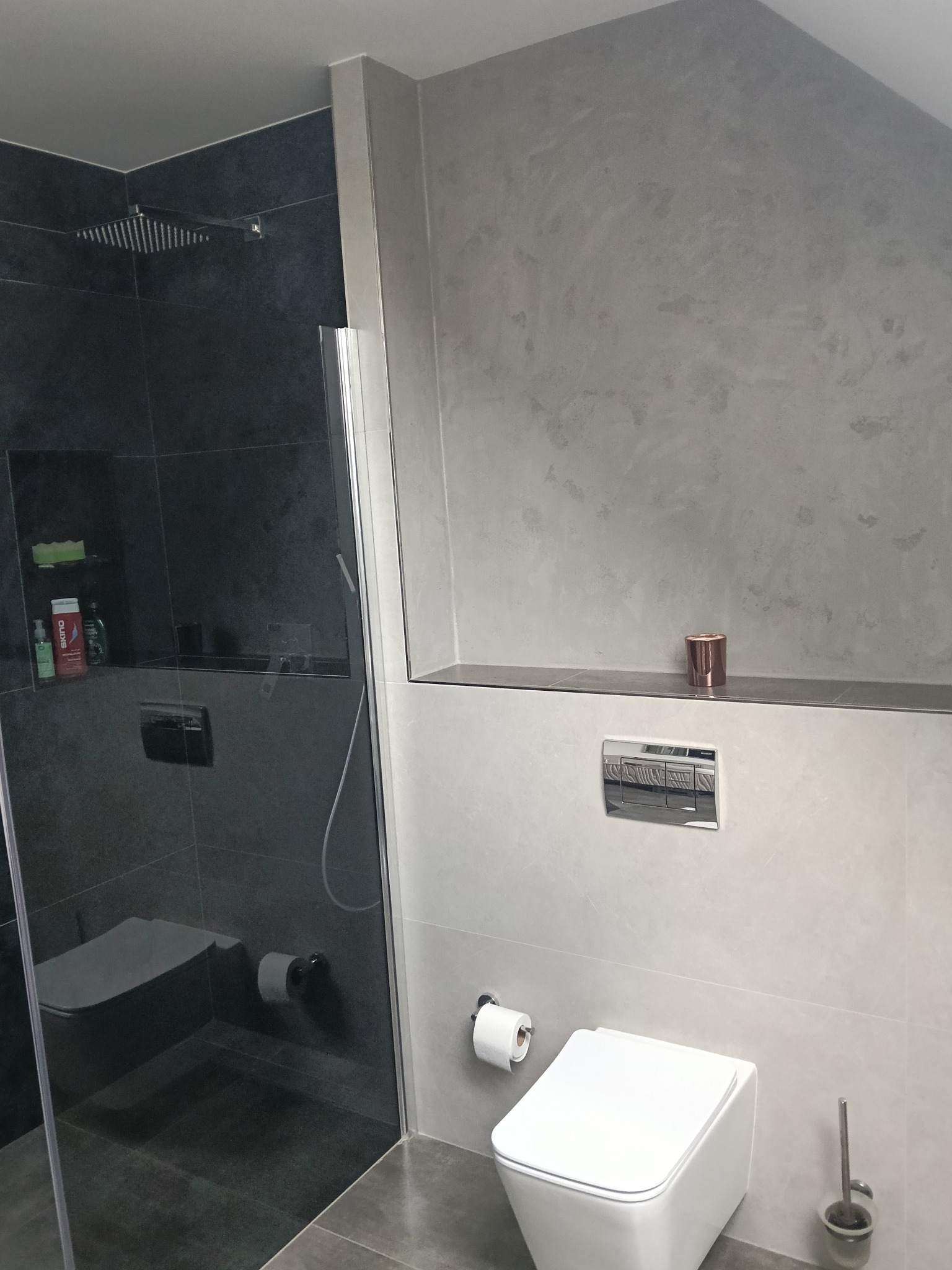 Nowoczesna łazienka w szarościach: kabina prysznicowa typu walk-in z ciemnymi płytkami, podtynkowa toaleta i dekoracyjna półka z akcentem miedzianym.