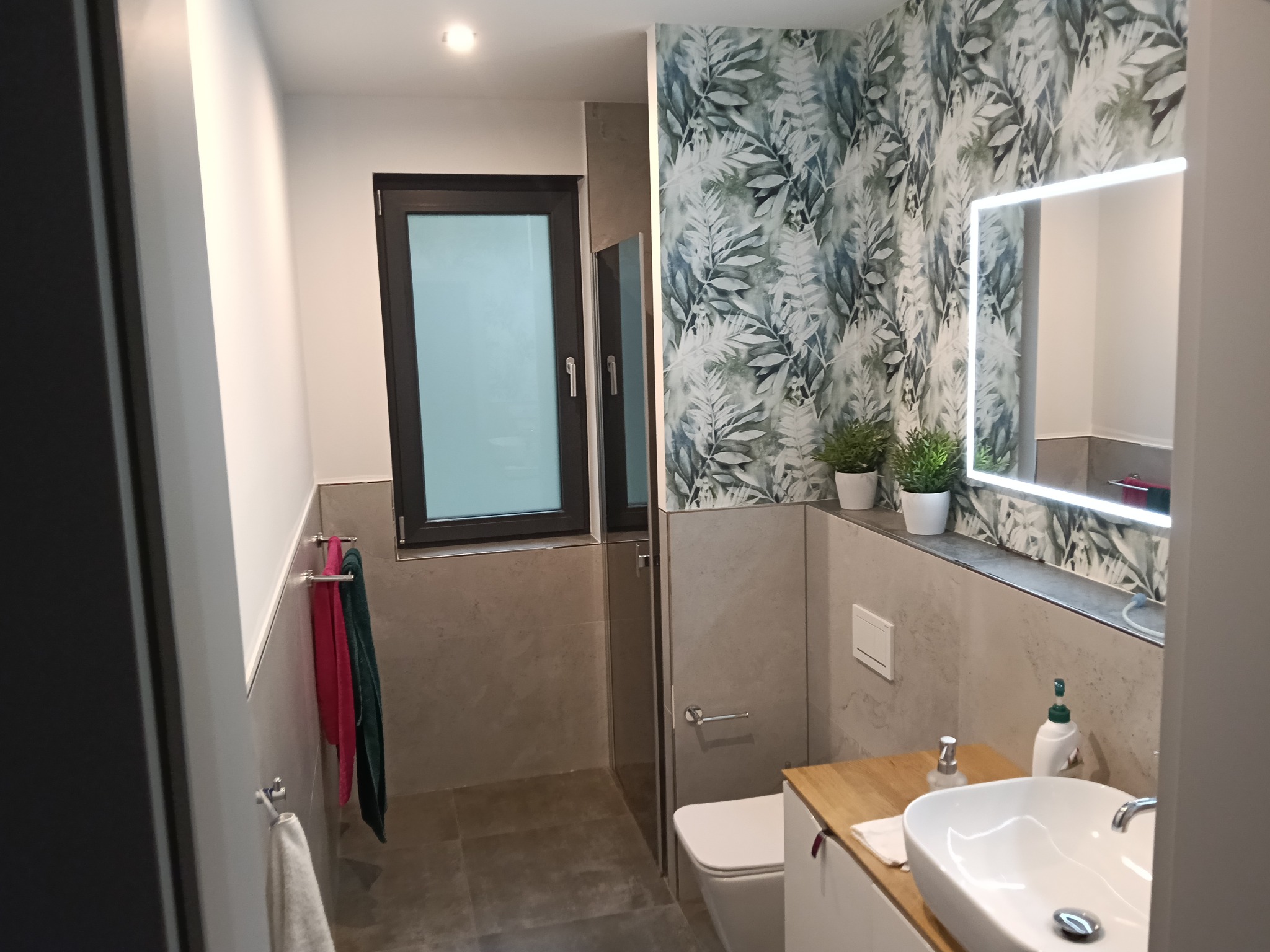 Nowoczesna łazienka po remoncie: biała umywalka, podświetlane lustro, roślinny motyw na ścianie, szare płytki i czarne okno. Minimalistyczny design.