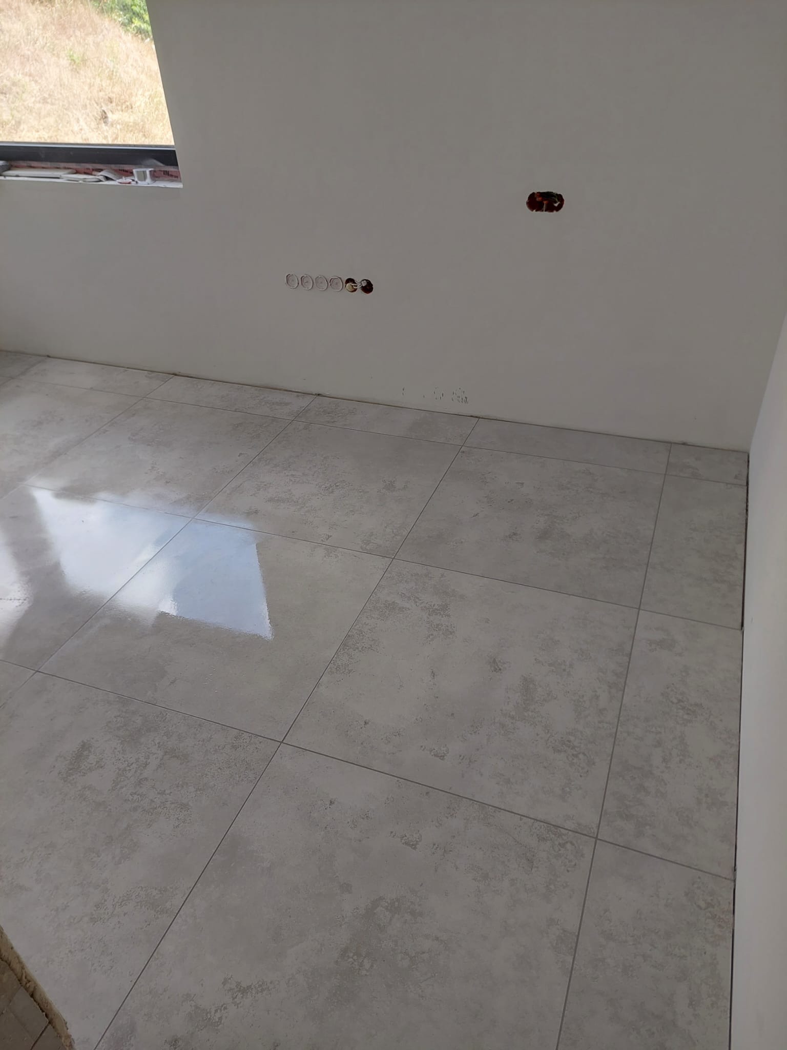 Jasnoszara, matowa podłoga z płytek w nowym wnętrzu. Widoczne gniazdka elektryczne na białej ścianie i okno z widokiem na zewnątrz. Minimalistyczny styl wykończenia.